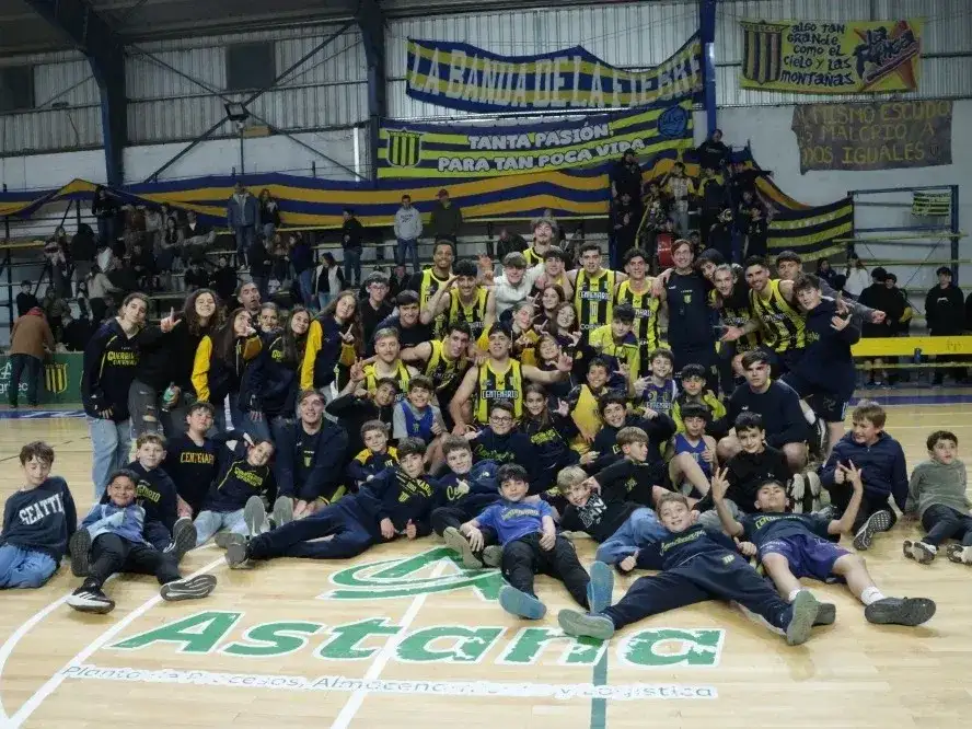 El plantel de básquet de Centenario celebra junto a sus hinchas y colaboradores tras el triunfo ante Rocamora por 88 a 66. El equipo posa en el campo de juego del estadio, rodeado de banderas y el apoyo de su gente en un inicio de torneo ideal.
