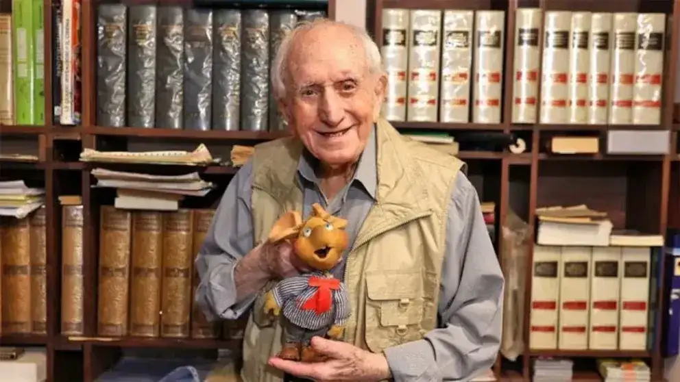 El actor italiano Peppino Mazzullo sostiene una figura del personaje Topo Gigio frente a una estantería llena de libros.