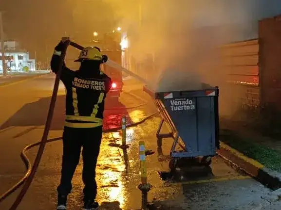 Personal de Bomberos Voluntarios de Venado Tuerto trabaja intensamente para sofocar un incendio desatado en un volquete de basura ubicado en la vía pública, utilizando una manguera de alta presión para extinguir las llamas y el humo generado.